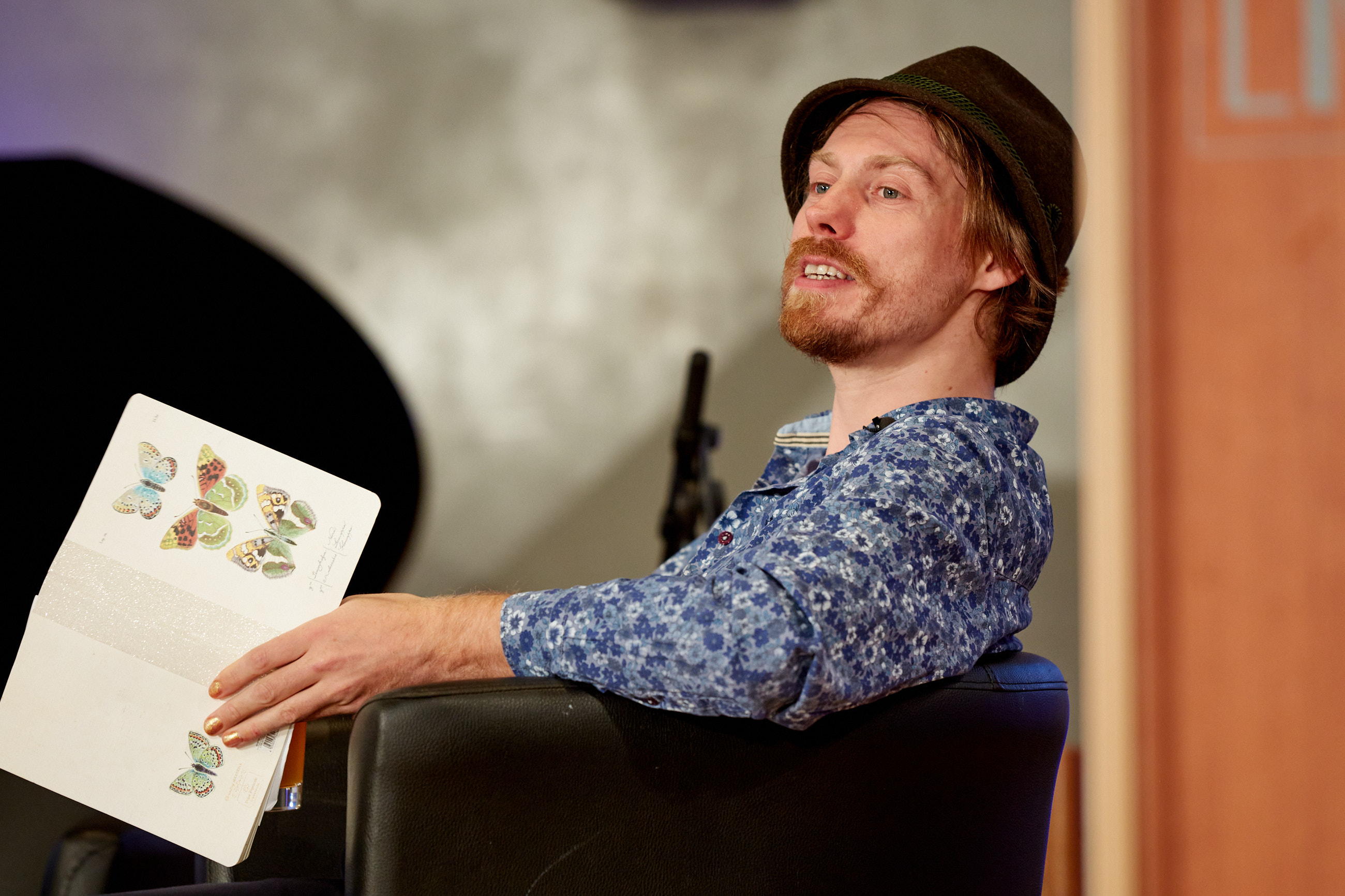Junger Mann mit Bart und Tirolerhut hat ein Buch über Schmetterlinge in der Hand und spricht